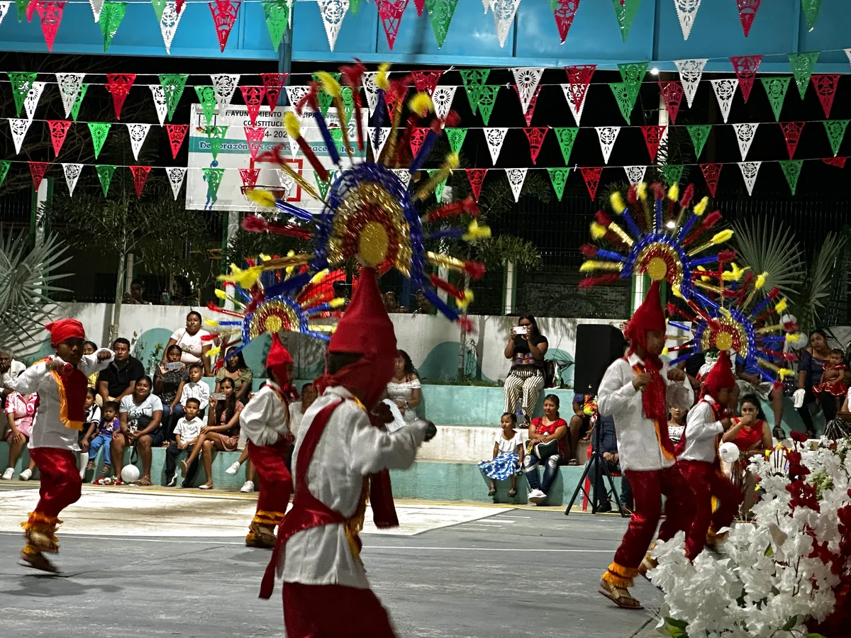 Danzantes con penachos de listones multicolor en forma de sol y traje rojo y blanco, junto a un arreglo de flores blancas.