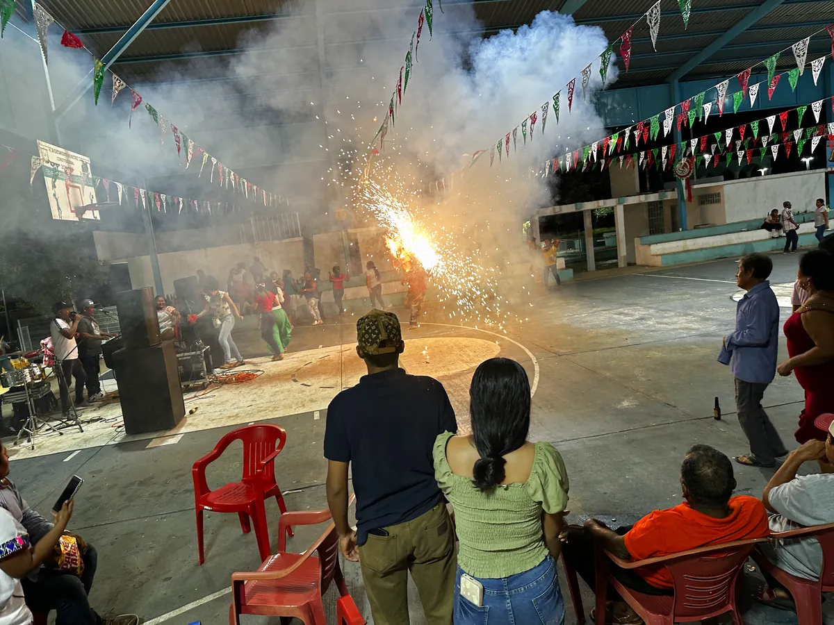 Un torito de fuegos artificiales encendido, lanzando chispas dentro del auditorio de la comunidad, con el público mirando alrededor.