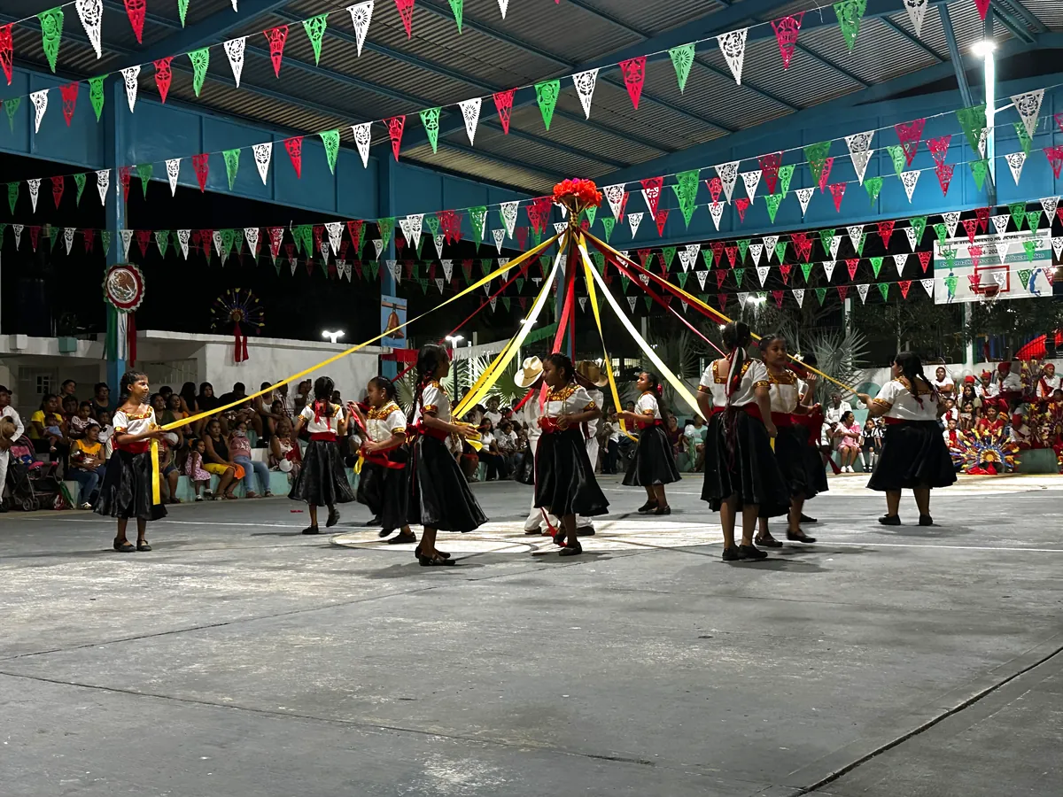 Niñas y niños bailando alrededor de un árbol de listones de colores bajo banderines de papel picado, en el auditorio de la comunidad.