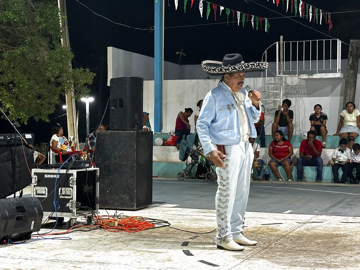 Un cantante de traje de charro azul claro canta con micrófono sobre el templete, con banderines de papel picado detrás.