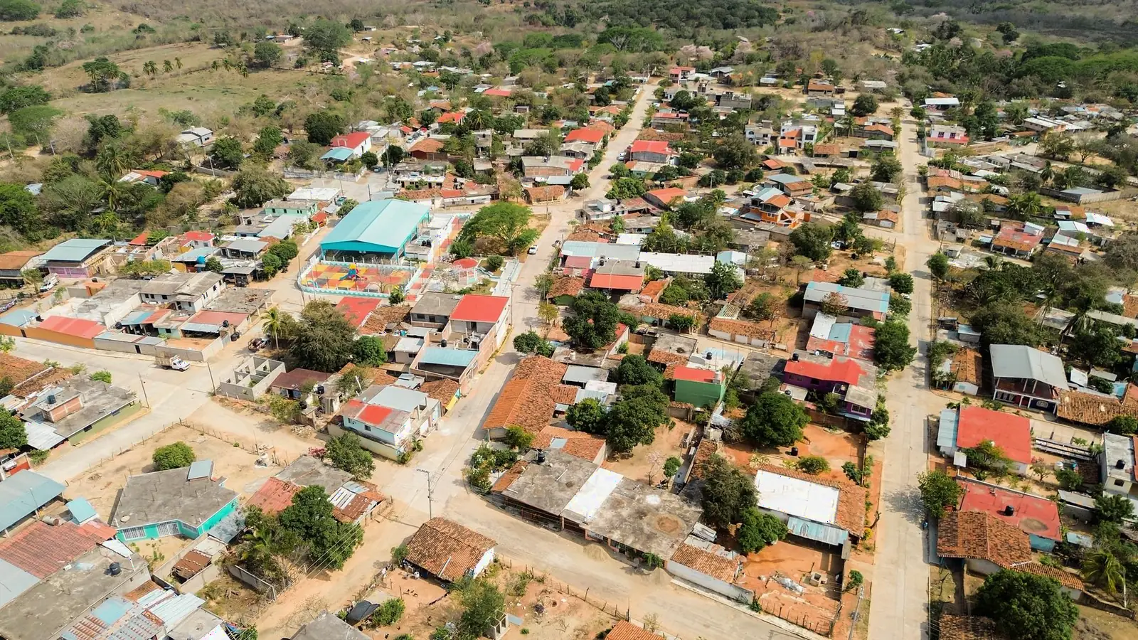 La plaza con su cancha de colores y el auditorio de techo turquesa de Agua Zarca, vistos desde el aire entre calles, techos de teja y monte.